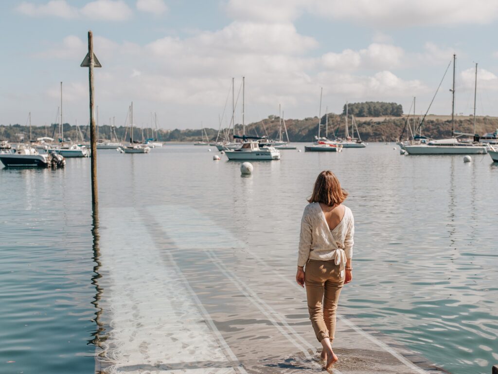 Une femme vue de dos s'avance vers l'eau de l'estuaire de la Rance en descendant un ponton à Pleurtuit, en Bretagne. Devant elle, des voiliers sont attachés à leurs bouées. Sur l'autre rive, la côte boisée de l'estuaire laisse entrevoir la rive droite de la Rance.