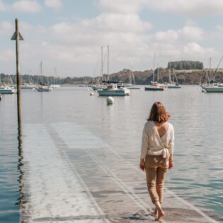 Une femme vue de dos s'avance vers l'eau de l'estuaire de la Rance en descendant un ponton à Pleurtuit, en Bretagne. Devant elle, des voiliers sont attachés à leurs bouées. Sur l'autre rive, la côte boisée de l'estuaire laisse entrevoir la rive droite de la Rance.