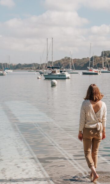 Une femme vue de dos s'avance vers l'eau de l'estuaire de la Rance en descendant un ponton à Pleurtuit, en Bretagne. Devant elle, des voiliers sont attachés à leurs bouées. Sur l'autre rive, la côte boisée de l'estuaire laisse entrevoir la rive droite de la Rance.