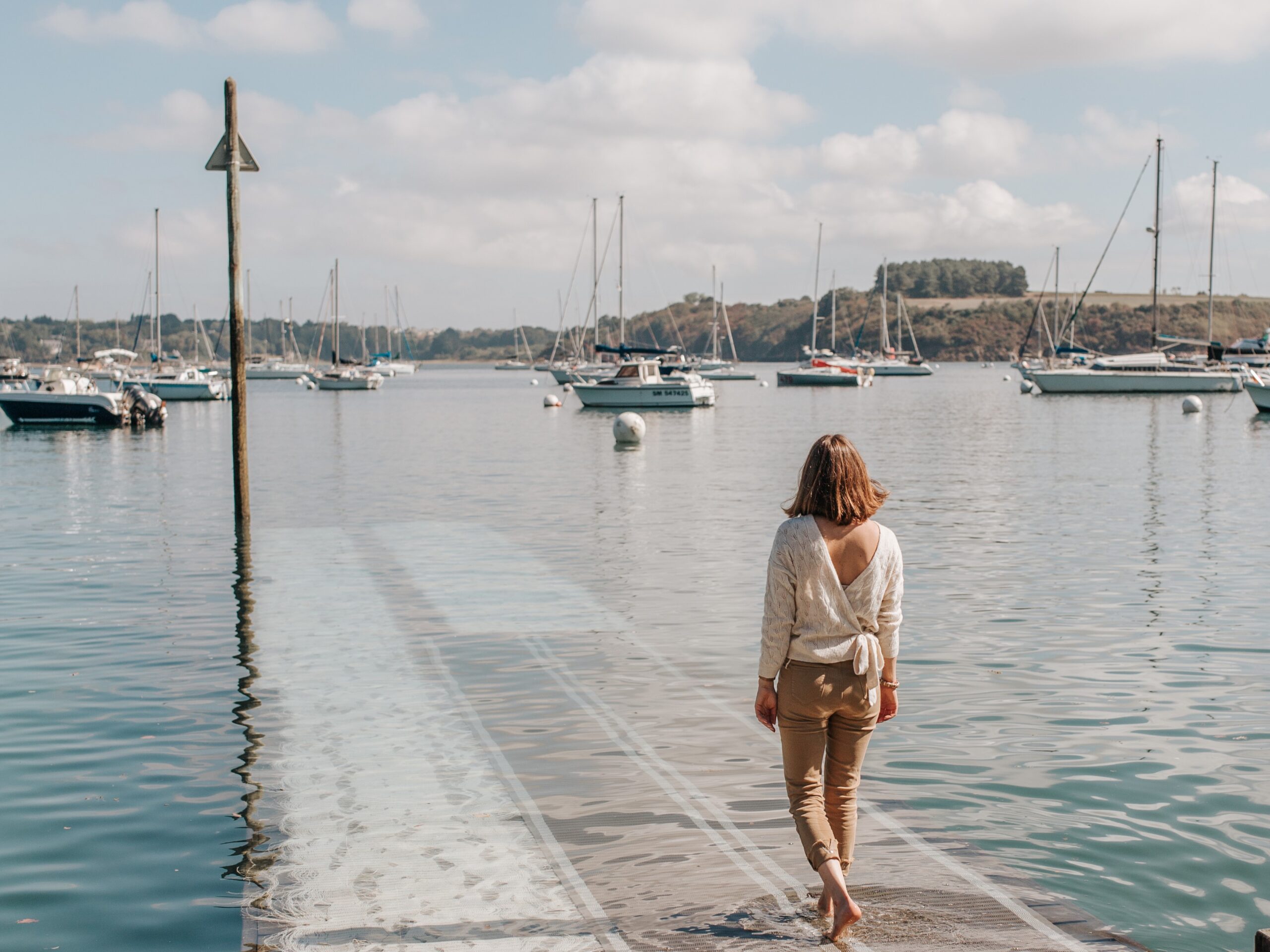 Une femme vue de dos s'avance vers l'eau de l'estuaire de la Rance en descendant un ponton. Devant elle, des voiliers sont attachés à leurs bouées. Sur l'autre rive, la côte boisée de l'estuaire laisse entrevoir la rive droite de la Rance.