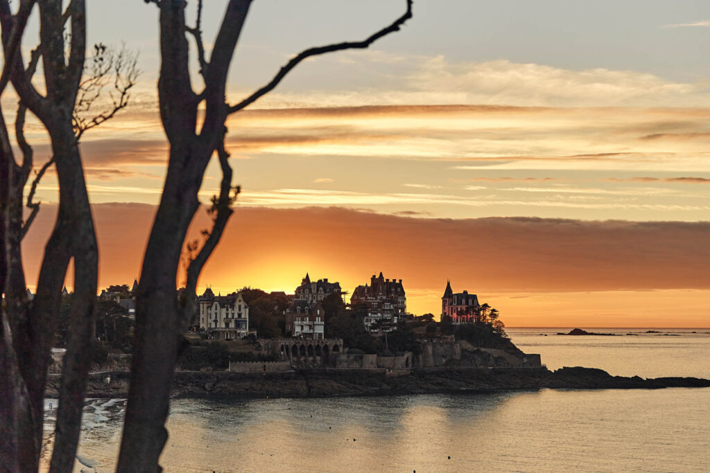 Les branches d'un arbre laissent entrevoir l'un des plus beaux Couchers de soleil de la Côte d'Émeraude : derrière la Pointe De La Malouine à Dinard, en Bretagne. Les villas Belle-Epoque situées sur la Pointe dominent la mer.