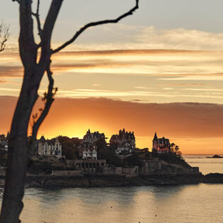 Les branches d'un arbre laissent entrevoir un Coucher de soleil aux tons jaune derrière la Pointe De La Malouine à Dinard, en Bretagne. Les villas Belle-Epoque situées sur la Pointe dominent la mer.