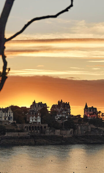 Les branches d'un arbre laissent entrevoir un Coucher de soleil aux tons jaune derrière la Pointe De La Malouine à Dinard, en Bretagne. Les villas Belle-Epoque situées sur la Pointe dominent la mer.