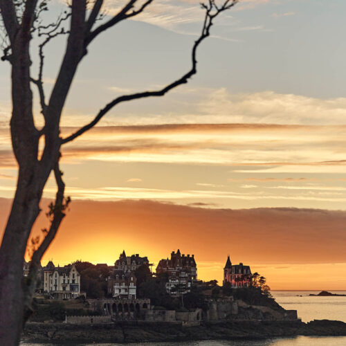 Les branches d'un arbre laissent entrevoir un Coucher de soleil aux tons jaune derrière la Pointe De La Malouine à Dinard, en Bretagne. Les villas Belle-Epoque situées sur la Pointe dominent la mer.