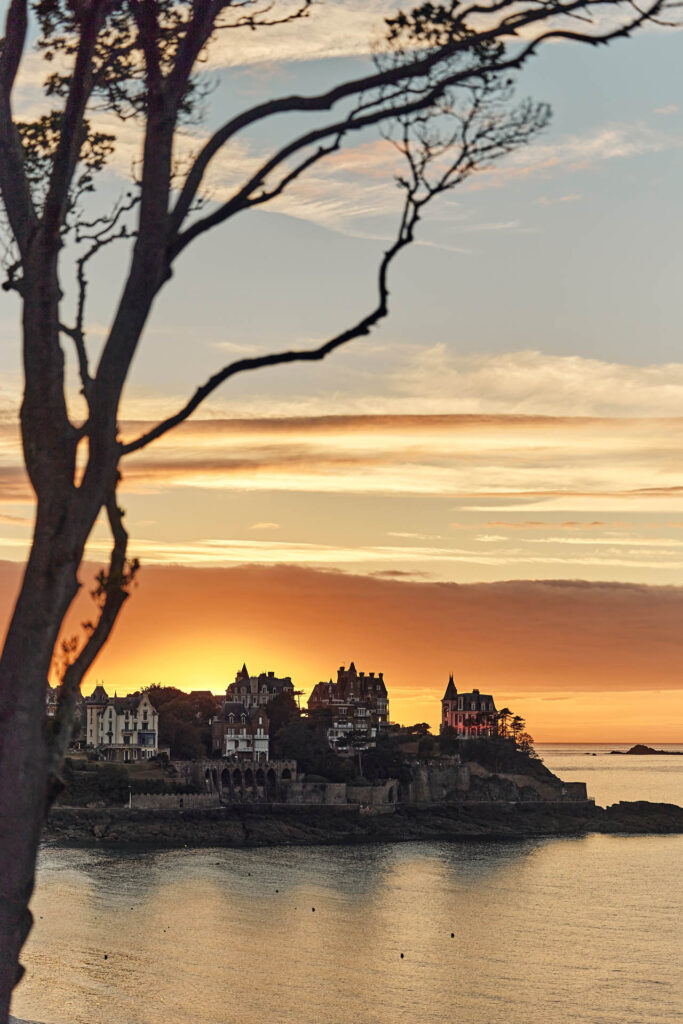 Les branches d'un arbre laissent entrevoir un Coucher de soleil aux tons jaune derrière la Pointe De La Malouine à Dinard, en Bretagne. Les villas Belle-Epoque situées sur la Pointe dominent la mer.