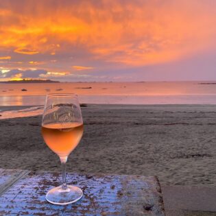 Coucher De Soleil Lancieux Sur Plage De Saint Sieu. Un verre de vin rosé est posé sur une table en bois à Kalypso La Cabane, restaurant de plage. La couleur du ciel ressemble à celle du verre de vin rosé.