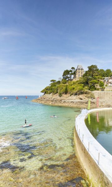 Dinard Pointe Du Moulinet, piscine d'eau de mer au bord de la plage de l'Ecluse. Les amateurs de sports nautiques pratiquent le stand-up paddle sur l'eau calme de la Manche. La villa Saint-Germain en arrière plan entourée de végétation.