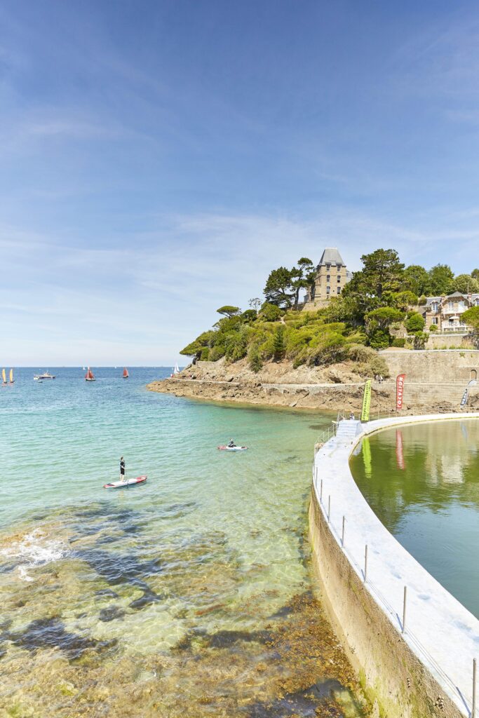 Dinard Pointe Du Moulinet, piscine d'eau de mer au bord de la plage de l'Ecluse. Les amateurs de sports nautiques pratiquent le stand-up paddle sur l'eau calme de la Manche. La villa Saint-Germain en arrière plan entourée de végétation.