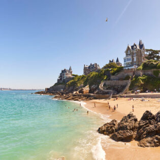 Les villas Belle-Epoque sur la pointe de la Malouine à Dinard en Bretagne. Des baigneurs sont dans l'eau turquoise de la Manche et sur le sable fin de la plage. Le soleil brille sur un goéland qui passe au dessus des villas.