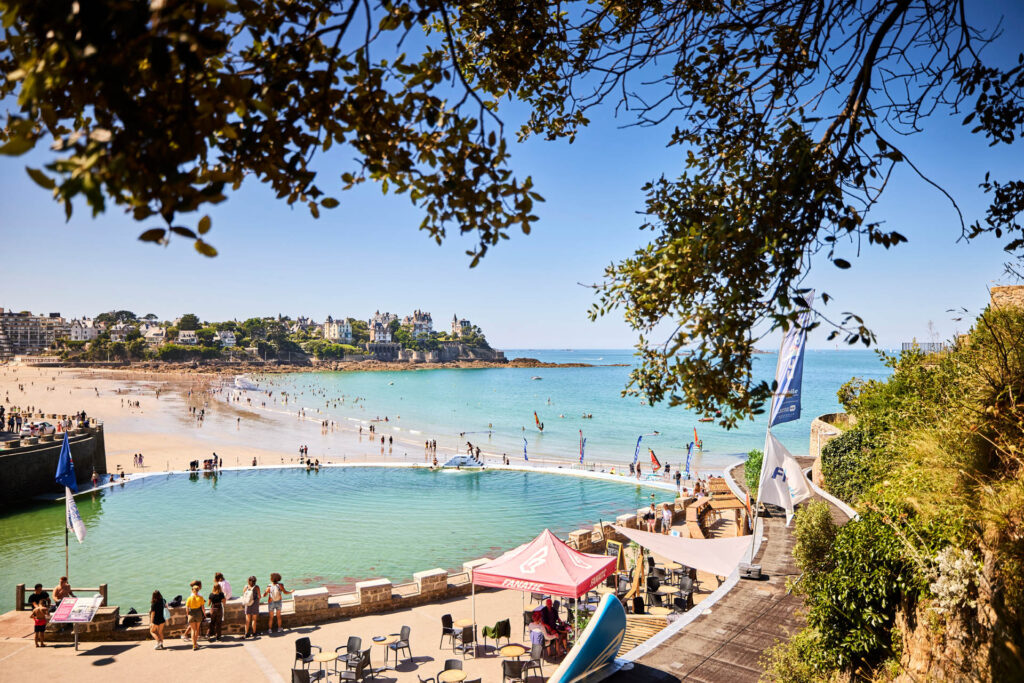 Vue en hauteur de la Piscine d'eau de mer située à Dinard au bord de la plage. De l'autre côté de la plage, les villas de la Pointe de la Malouine se trouve au dessus du sentier côtier. Sur la plage, des personnes se baignent et au bord de la piscine, le bar accueille des clients installés au soleil.