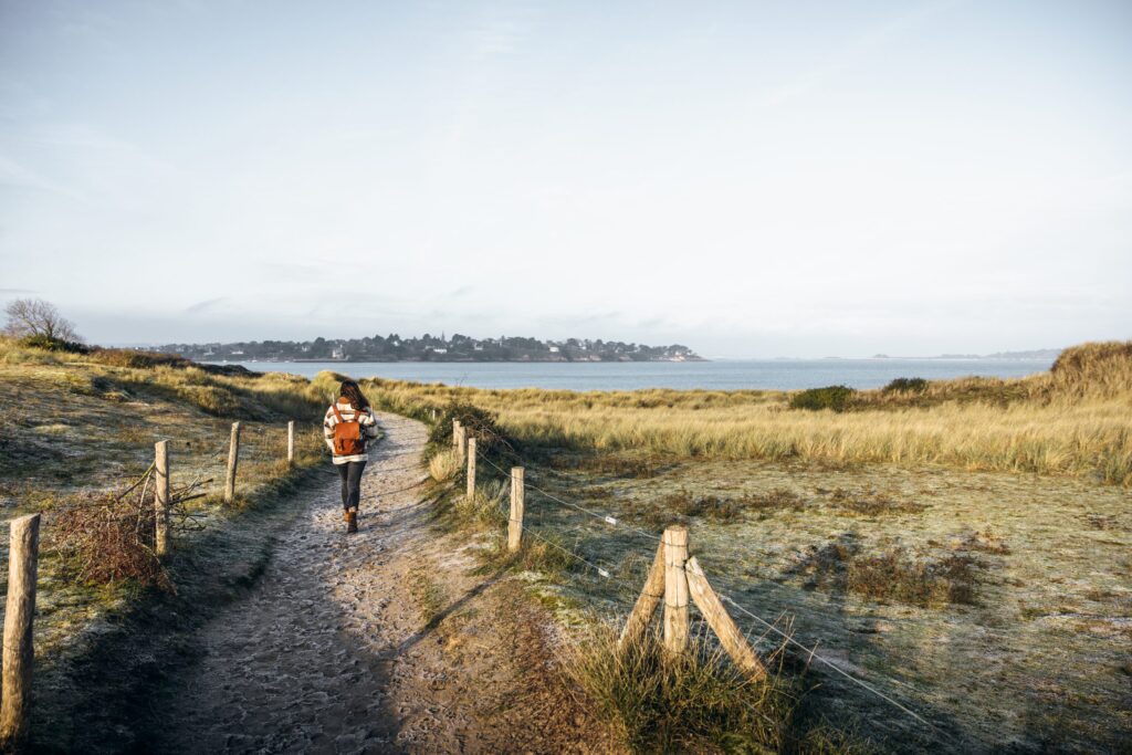 Photo en hiver du Tertre Corlieu et la plage des Briantais à Lancieux. Une femme vue de dos marche sur un petit chemin aux reflets blancs du givre. Le chemin est bien délimité et des deux côtés, les dunes sableuses s'étendent jusqu'à la mer. De l'autre côté de la baie de Lancieux se trouve le village de Saint-Jacut de la Mer.