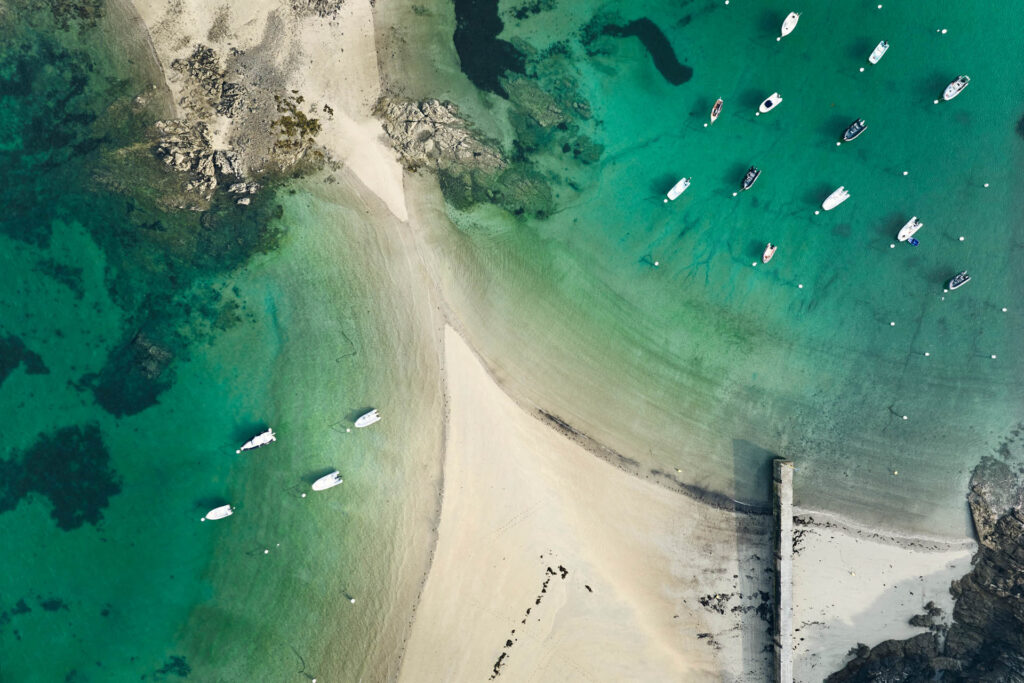 Tombolo de la Plage De L'islet à Lancieux vu du ciel. Des deux côtés du tombolo, la mer couleur émeraude et des bateaux accrochés à leurs bouées. Un ponton en pierre s'avance vers la mer.