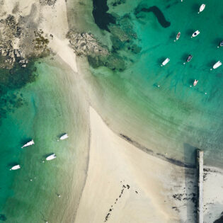 Tombolo de la Plage De L'islet à Lancieux vu du ciel. Des deux côtés du tombolo, la mer couleur émeraude et des bateaux accrochés à leurs bouées. Un ponton en pierre s'avance vers la mer.