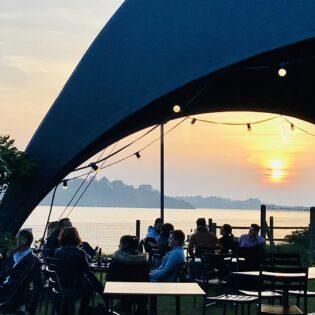 Coucher de soleil derrière la grande tente installée sur la terrasse du bar Le Dôme By Camping Port Blanc à Dinard, le long de la plage de Port Blanc. Des personnes sont attablées et discutent.