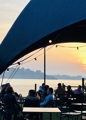 Coucher de soleil derrière la grande tente installée sur la terrasse du bar Le Dôme By Camping Port Blanc à Dinard, le long de la plage de Port Blanc. Des personnes sont attablées et discutent.