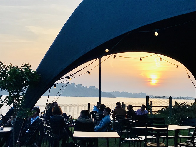 Pour admirer un coucher de soleil sur la Côte d'Emeraude, installez-vous sur la terrasse du bar Le Dôme By Camping Port Blanc à Dinard, le long de la plage de Port Blanc. Des personnes sont attablées et discutent.