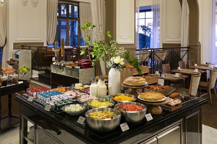 Au coeur de la salle de restaurant du Grand Hotel Barriere de Dinard, le buffet du brunch est copieux et varié. Un bouquet de fleur vient décorer cette table de buffet. Autour, des tables sont prêtes à accueillir les clients.