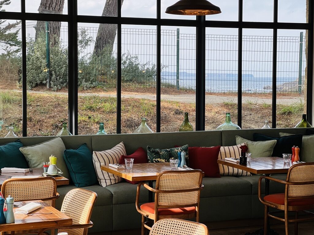 Tables, chaises et banquettes confortables avec des coussins colorés, disposés le long de grandes baies vitrées au Restaurant Du Nessay à Saint-Briac-sur-Mer. De l'intérieur du restaurant, le panorama s'ouvre sur la mer et le Cap Fréhel.