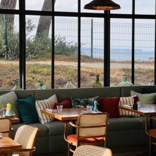 Tables, chaises et banquettes confortables avec des coussins colorés, disposés le long de grandes baies vitrées au Restaurant Du Nessay à Saint-Briac-sur-Mer. De l'intérieur du restaurant, le panorama s'ouvre sur la mer et le Cap Fréhel.