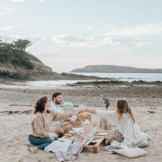 Trois personnes ont organisé un picnic sur la plage du Perron à Saint-Briac-sur-Mer au coucher du soleil. En arrière plan, l'île Agot se trouve de l'autre côté d'un bras de mer.