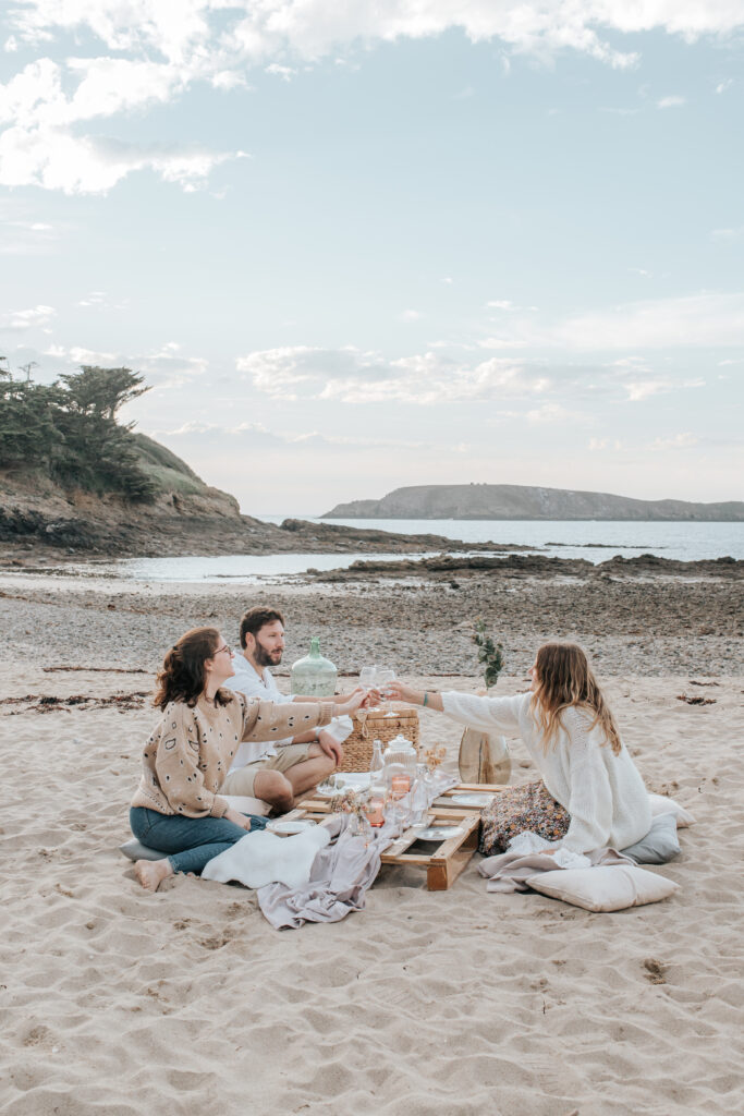 Trois personnes ont organisé un picnic sur la plage du Perron à Saint-Briac-sur-Mer au coucher du soleil. En arrière plan, l'île Agot se trouve de l'autre côté d'un bras de mer.