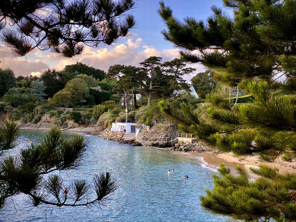 Plage De La Petite Salinette, une des plus jolies criques de la Côte d'Émeraude, située à Saint Briac Sur Mer, sur la Côte d'Emeraude en Bretagne. Entre les pins maritimes, des baigneurs profitent d'une baignade dans la mer en début de soirée. La marée haute recouvre presque toute la plage. Le cabanon blanc du Yacht Club se trouve entre la plage et la végétation en hauteur.