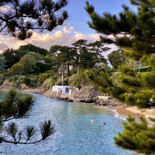 Plage De La Petite Salinette, une des plus jolies criques de la Côte d'Émeraude, située à Saint Briac Sur Mer, sur la Côte d'Emeraude en Bretagne. Entre les pins maritimes, des baigneurs profitent d'une baignade dans la mer en début de soirée. La marée haute recouvre presque toute la plage. Le cabanon blanc du Yacht Club se trouve entre la plage et la végétation en hauteur.