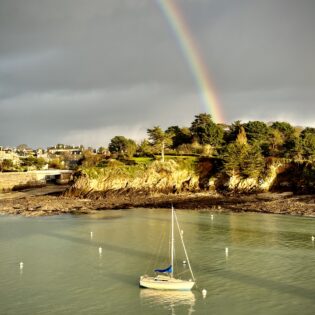 Un Arc-en-ciel apparaît à Saint Briac sur Mer, au milieu d'un ciel gris et une eau vert émeraude. Un voilier se trouve au mouillage d'une eau paisible.