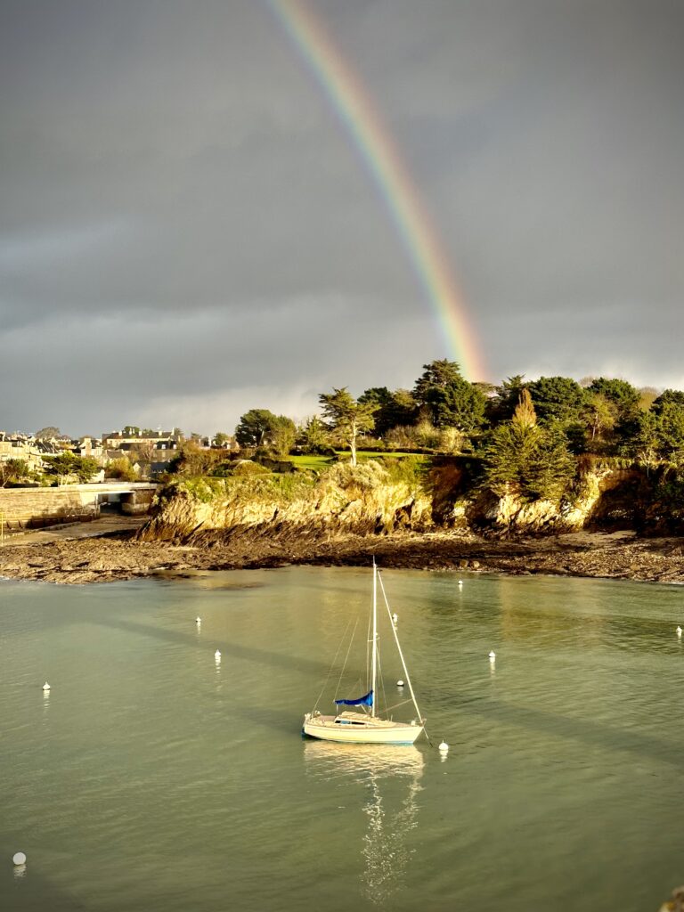 Un Arc-en-ciel apparaît à Saint Briac sur Mer, au milieu d'un ciel gris et une eau vert émeraude. Un voilier se trouve au mouillage d'une eau paisible.