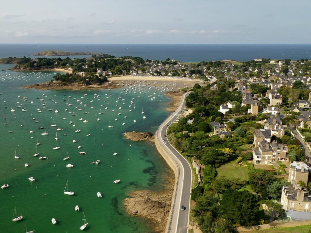 Vue aérienne du Balcon d'Emeraude à Saint-Briac-sur-Mer en Bretagne. Les voiliers sont au mouillage au port de plaisance, situé entre des villas balnéaires et le Chateau du Nessay. En arrière plan, la Manche s'étend à l'horizon. Une plage concave borde le port de plaisance.