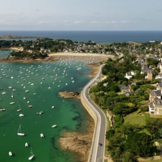 Vue aérienne du Balcon d'Emeraude à Saint-Briac-sur-Mer en Bretagne. Les voiliers sont au mouillage au port de plaisance, situé entre des villas balnéaires et le Chateau du Nessay. En arrière plan, la Manche s'étend à l'horizon. Une plage concave borde le port de plaisance.