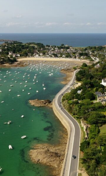 Vue aérienne du Balcon d'Emeraude à Saint-Briac-sur-Mer en Bretagne. Les voiliers sont au mouillage au port de plaisance, situé entre des villas balnéaires et le Chateau du Nessay. En arrière plan, la Manche s'étend à l'horizon. Une plage concave borde le port de plaisance.