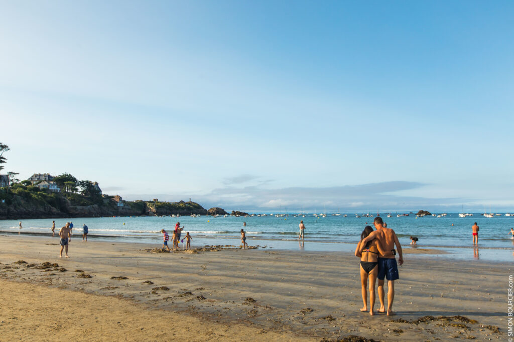 Un couple se promène bras dessus bras dessous sur la Grande Plage de Saint-Lunaire au cœur de l'été. Un grand ciel bleu offre la possibilité aux baigneurs et aux famille de profiter des joies de la plage : châteaux de sable, baignades, etc. Cette plage est bordée par la Pointe du Décollé, et notamment sa croix monumentale située à son extrémité.