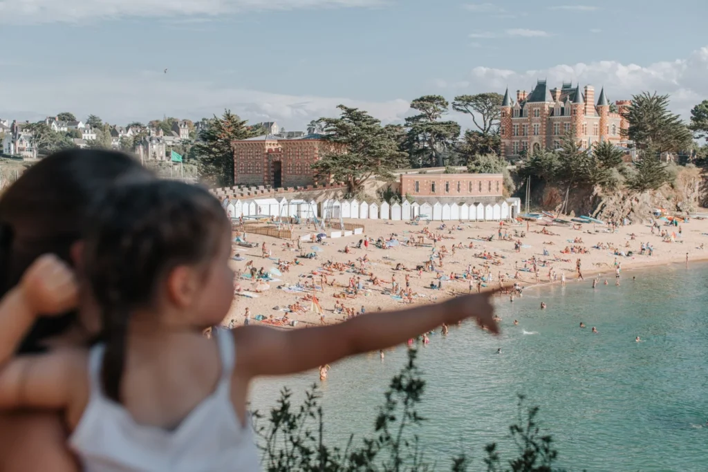 Une petite fille pointe du doigt la plage de la Grande Salinette à Saint-Briac. En arrière-plan, on aperçoit l'hôtel du Nessay, baigné par la lumière du soleil.