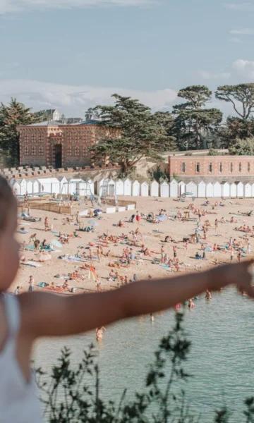 Une petite fille pointe du doigt la plage de la Grande Salinette à Saint-Briac. En arrière-plan, on aperçoit l'hôtel du Nessay, baigné par la lumière du soleil.