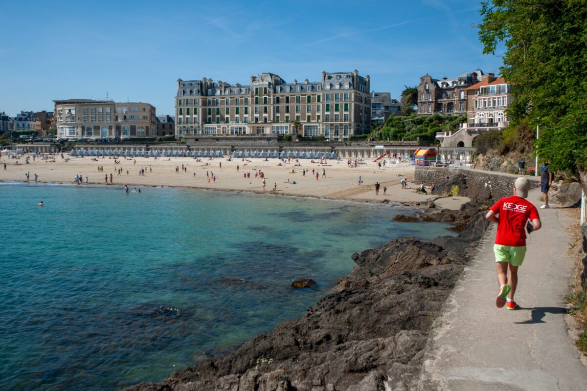 Un homme en sweat rouge court sur le gr34 de Dinard. Il s'agit de la pointe de la malouine, on aperçoit en arrière plan la plage de l'Écluse et le Royal.