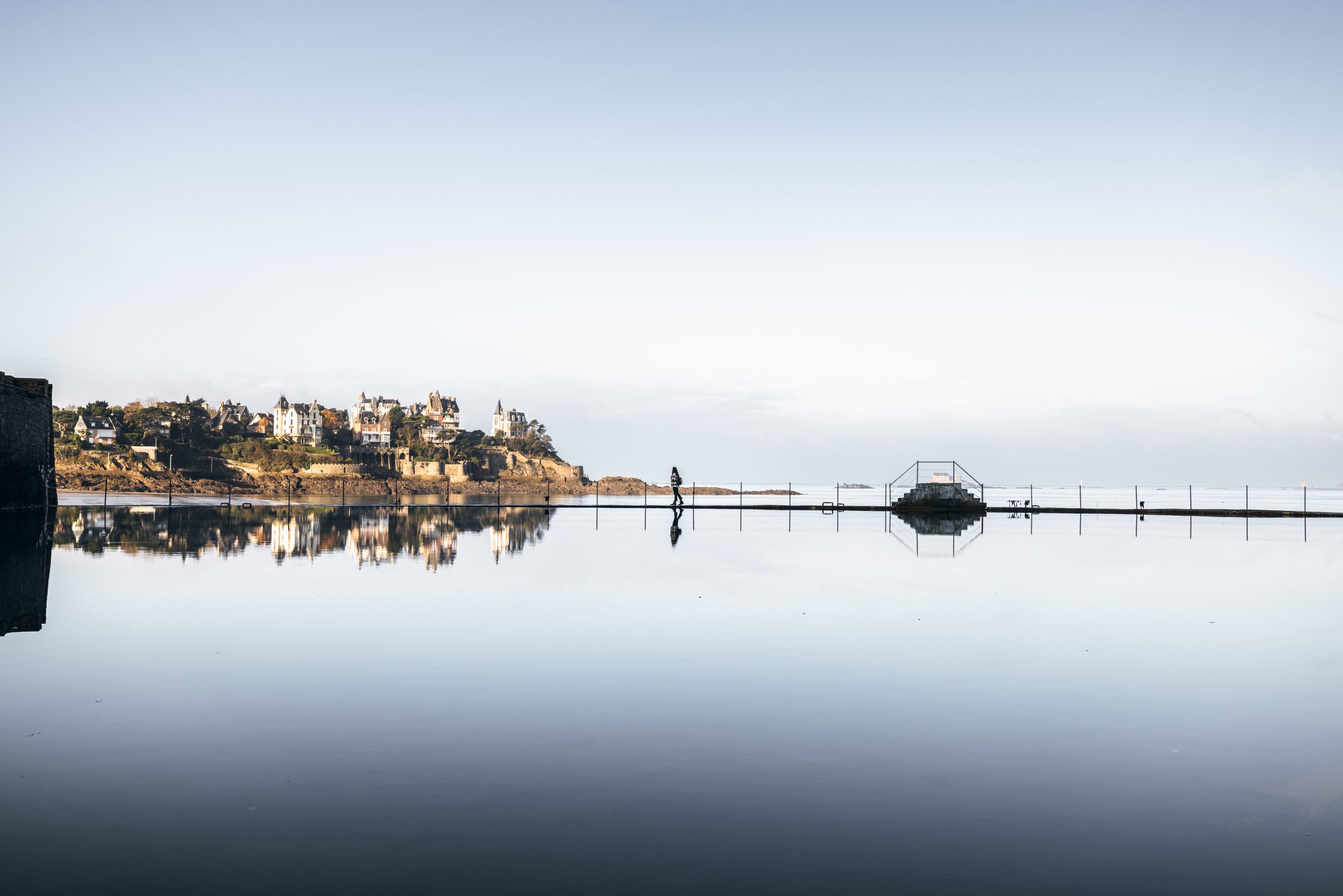 Une jeune fille marche au bord de la piscine à Dinard durant l'hiver. Derrière elle, des villas élégantes servent de toile de fond à la scène paisible. La piscine est tranquille et reflète les nuances grises du ciel hivernal.