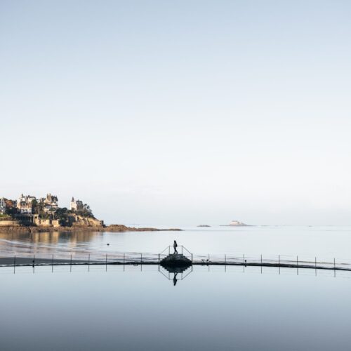 Une jeune fille marche au bord de la piscine à Dinard durant l'hiver. Derrière elle, des villas élégantes servent de toile de fond à la scène paisible. La piscine est tranquille et reflète les nuances grises du ciel hivernal.