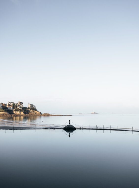 Une jeune fille marche au bord de la piscine à Dinard durant l'hiver. Derrière elle, des villas élégantes servent de toile de fond à la scène paisible. La piscine est tranquille et reflète les nuances grises du ciel hivernal.