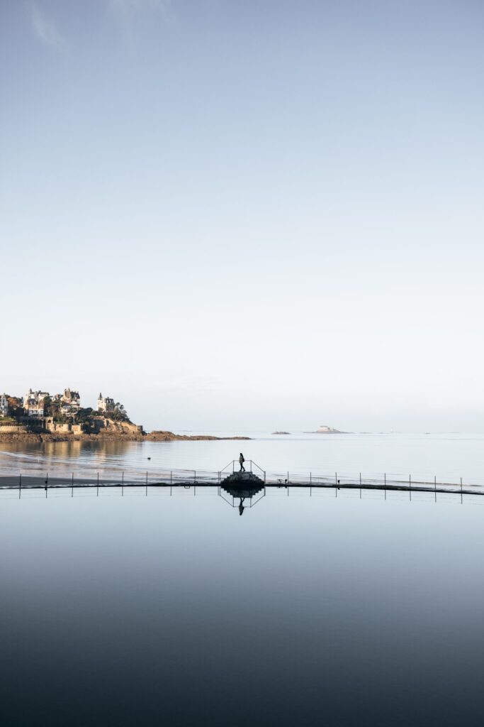 Une jeune fille marche au bord de la piscine à Dinard durant l'hiver. Derrière elle, des villas élégantes servent de toile de fond à la scène paisible. La piscine est tranquille et reflète les nuances grises du ciel hivernal.