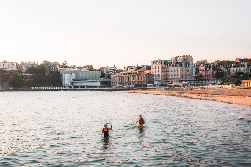 Sur la Plage de l'Écluse à Dinard, un couple de retraités se baigne dans la mer sous une lumière douce de début de journée. La lumière matinale teinte l'environnement de couleurs pastel, et le calme de la mer ajoute une sérénité à l'atmosphère. Le couple, en pleine baignade, semble apprécier ce moment paisible, entouré par la beauté naturelle de la plage au lever du soleil.