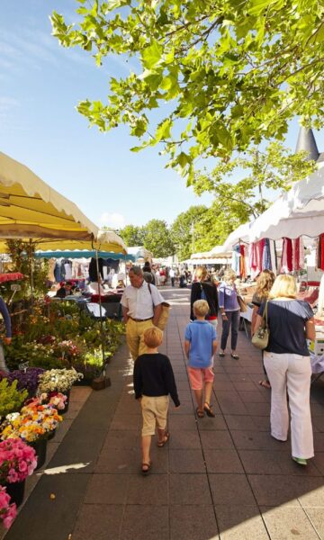 Allée du marché de Dinard. Des passants font leurs emplettes sous le soleil au marché de Dinard. L'un des plus grands de la Côte d'Émeraude, il a lieu le mardi, le jeudi et le samedi.