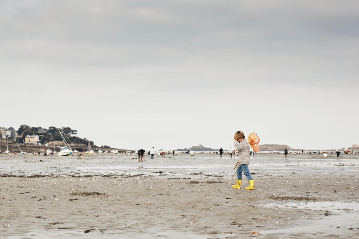 Une personne marchant sur la plage à marée basse près de Dinard, en Bretagne, France, recherche des coquillages et des crustacés dans un cadre pittoresque. L'individu, équipé d'un seau et d'un outil de fouille, profite de cette activité traditionnelle de pêche à pied, entouré par l'immensité de l'océan à l'horizon et les rochers émergeant du sable. Le ciel est légèrement nuageux, laissant filtrer la lumière douce du soleil couchant, ce qui ajoute une ambiance paisible et romantique à la scène.