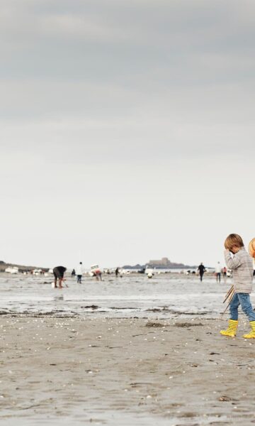 Un enfant marchant sur la plage à marée basse près de Dinard, en Bretagne, France, recherche des coquillages et des crustacés dans un cadre pittoresque. L'individu, équipé d'un seau et d'un outil de fouille, profite de cette activité traditionnelle de pêche à pied, entouré par l'immensité de l'océan à l'horizon et les rochers émergeant du sable. Le ciel est légèrement nuageux, laissant filtrer la lumière douce du soleil couchant, ce qui ajoute une ambiance paisible et romantique à la scène.