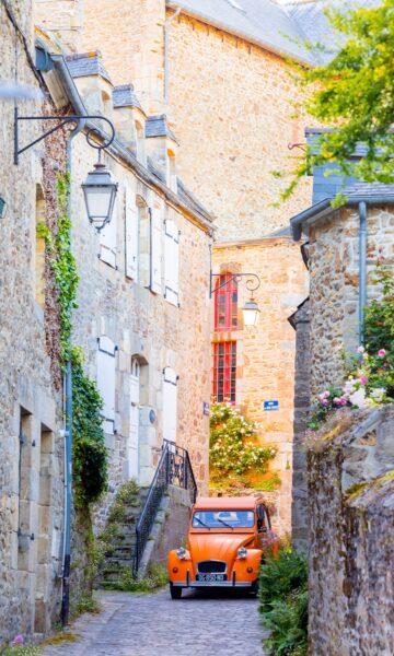 Une Citroën 2CV de couleur orange serpente à travers les ruelles étroites et pittoresques de Saint-Briac, offrant une touche de couleur vive contre le charme historique du village.