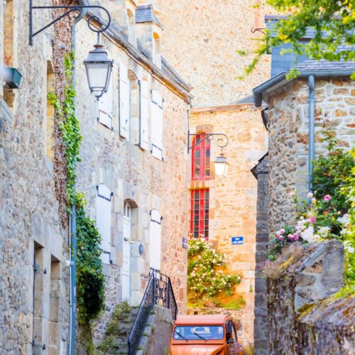 Une Citroën 2CV de couleur orange serpente à travers les ruelles étroites et pittoresques de Saint-Briac, offrant une touche de couleur vive contre le charme historique du village.