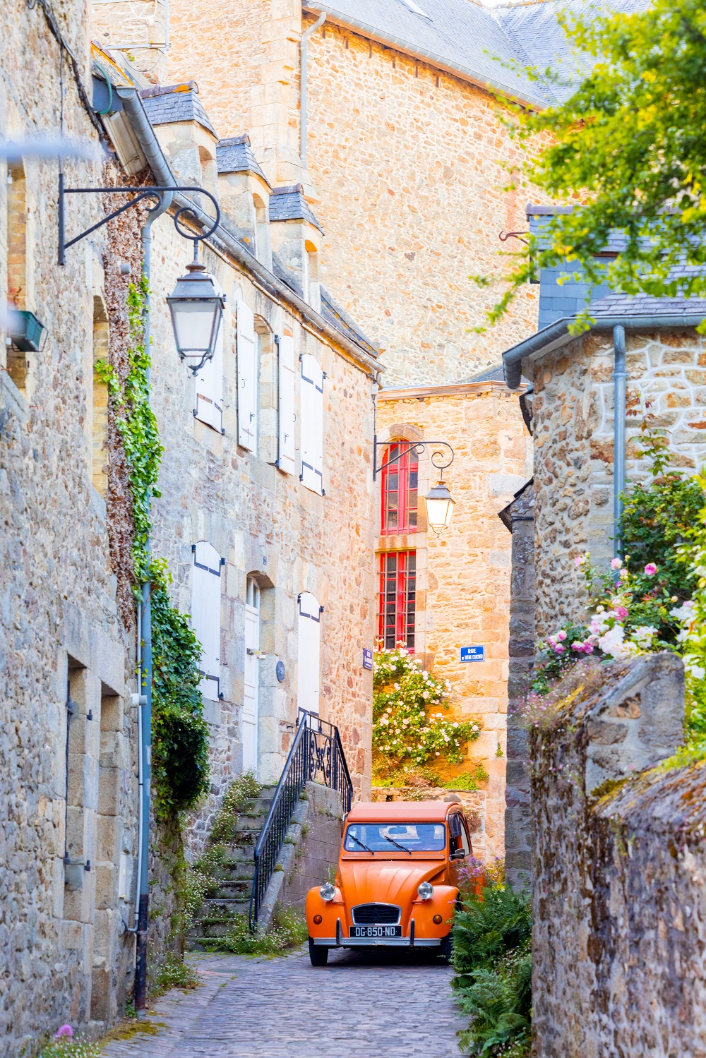 Une Citroën 2CV de couleur orange serpente à travers les ruelles étroites et pittoresques de Saint-Briac, offrant une touche de couleur vive contre le charme historique du village.