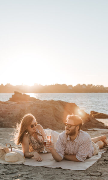 Scène romantique. Un couple allongé sur une fouta, buvant une coupe de champagne regarde le coucher du soleil à Lancieux sur la plage des Briantais