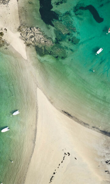 Vue aérienne de la Plage de L'islet à Lancieux. L'eau est bleu, vert émeraude. Des bateaux sont visibles.