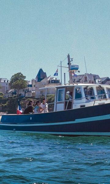 Le bateau de L'échappée Malouine navigue devant la Grande Plage de Saint-Lunaire et la Pointe du Décollé. Privatisez une croisière pour vos amis et votre famille sur la Côte d'Émeraude, pour découvrir la Baie de Saint-Malo en bateau !